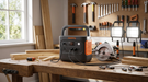 A Jackery Explorer 2000 v2 portable power station on a timber workbench in a home workshop, running a circular saw and two LED work lights, with a pegboard of hand tools behind.
