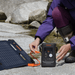 A person in outdoor gear connects an orange cable from a foldable Jackery solar panel to a Jackery portable power station on rocky ground by a stream, highlighting easy off-grid USB-C charging with Jackery products.