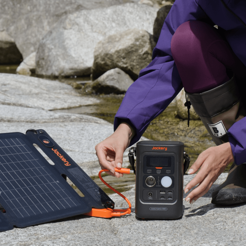 A person in outdoor gear connects an orange cable from a foldable Jackery solar panel to a Jackery portable power station on rocky ground by a stream, highlighting easy off-grid USB-C charging with Jackery products.