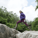 A person in a purple jacket, purple pants, and boots steps across large rocks outdoors, holding a compact Jackery portable power station. Green trees and a cloudy sky set the background scene.