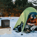 A man in an orange jacket and gray beanie sits on snow by a green tent amid trees. Next to him, a Jackery power station rests on a white storage box, offering reliable cold-weather protection and waterproof, dustproof performance.