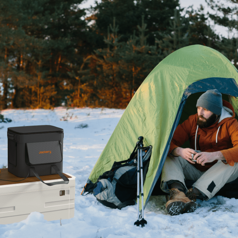A man in an orange jacket and gray beanie sits on snow by a green tent amid trees. Next to him, a Jackery power station rests on a white storage box, offering reliable cold-weather protection and waterproof, dustproof performance.