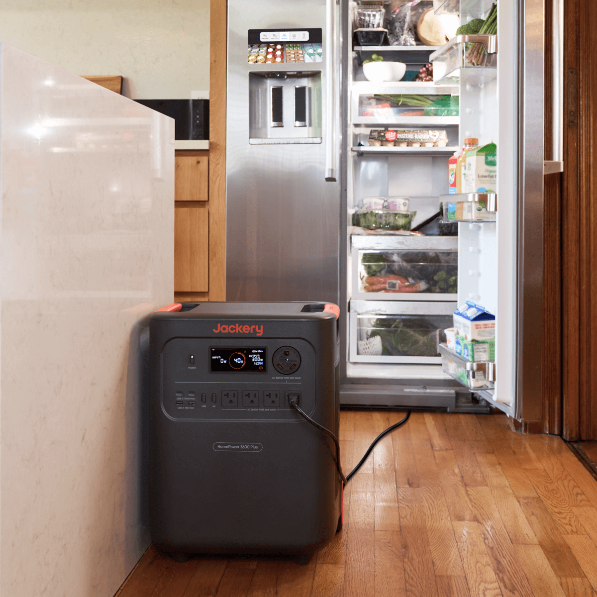 A Jackery portable power station sits on a wooden kitchen floor beside an open refrigerator stocked with food, ensuring your groceries and drinks stay fresh even during power outages.