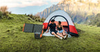 Two people relax on a blanket outside a red and white tent with mountains in the background. Beside them sits a Jackery portable solar generator and power station, ideal for charging devices during outdoor adventures.