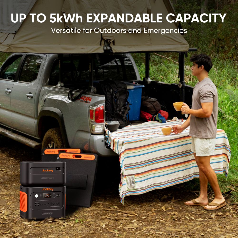 A man stands by a pickup truck with its tailgate down, covered by a striped cloth. Camping gear is arranged in the bed. Overlay text highlights the Jackery portable power station with up to 5kWh expandable capacity for outdoor use.