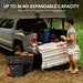 A man stands by a pickup truck with its tailgate down, covered by a striped cloth. Camping gear is arranged in the bed. Overlay text highlights the Jackery portable power station with up to 5kWh expandable capacity for outdoor use.