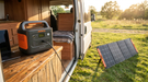 A Jackery Explorer 1000 v2 power station on a camper van worktop charging a laptop, with a cable running out through the open side door to a Jackery SolarSaga 200W solar panel standing on the grass in evening sun.