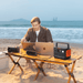 A person uses a Jackery E300v2 portable power station at a wooden beach table, powering a laptop, camera, speaker, and another laptop with the ocean and distant buildings in the background.