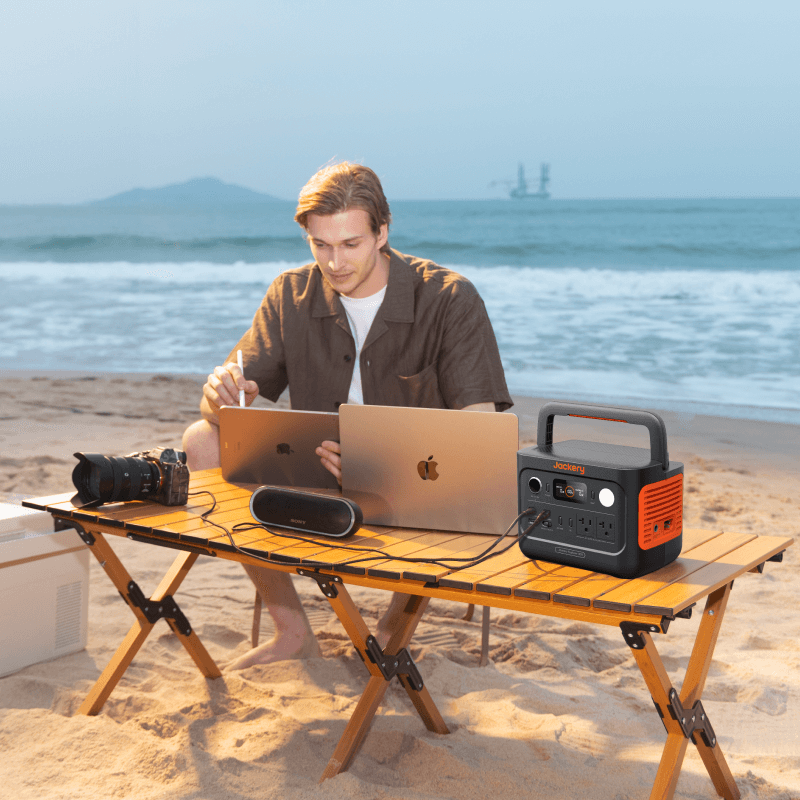 A person uses a Jackery E300v2 portable power station at a wooden beach table, powering a laptop, camera, speaker, and another laptop with the ocean and distant buildings in the background.