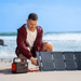 A person kneels on a sandy beach, connecting a Jackery portable power station with LiFePO4 battery to a solar panel. The ocean and blue sky are visible in the background.