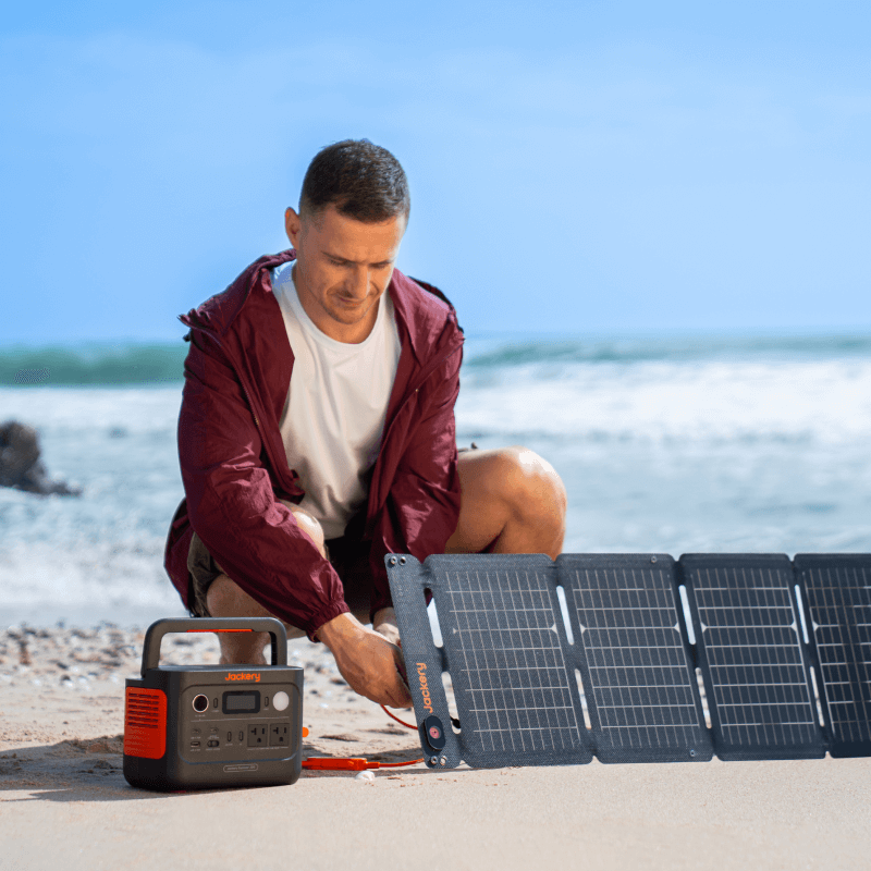 A person kneels on a sandy beach, connecting a Jackery portable power station with LiFePO4 battery to a solar panel. The ocean and blue sky are visible in the background.