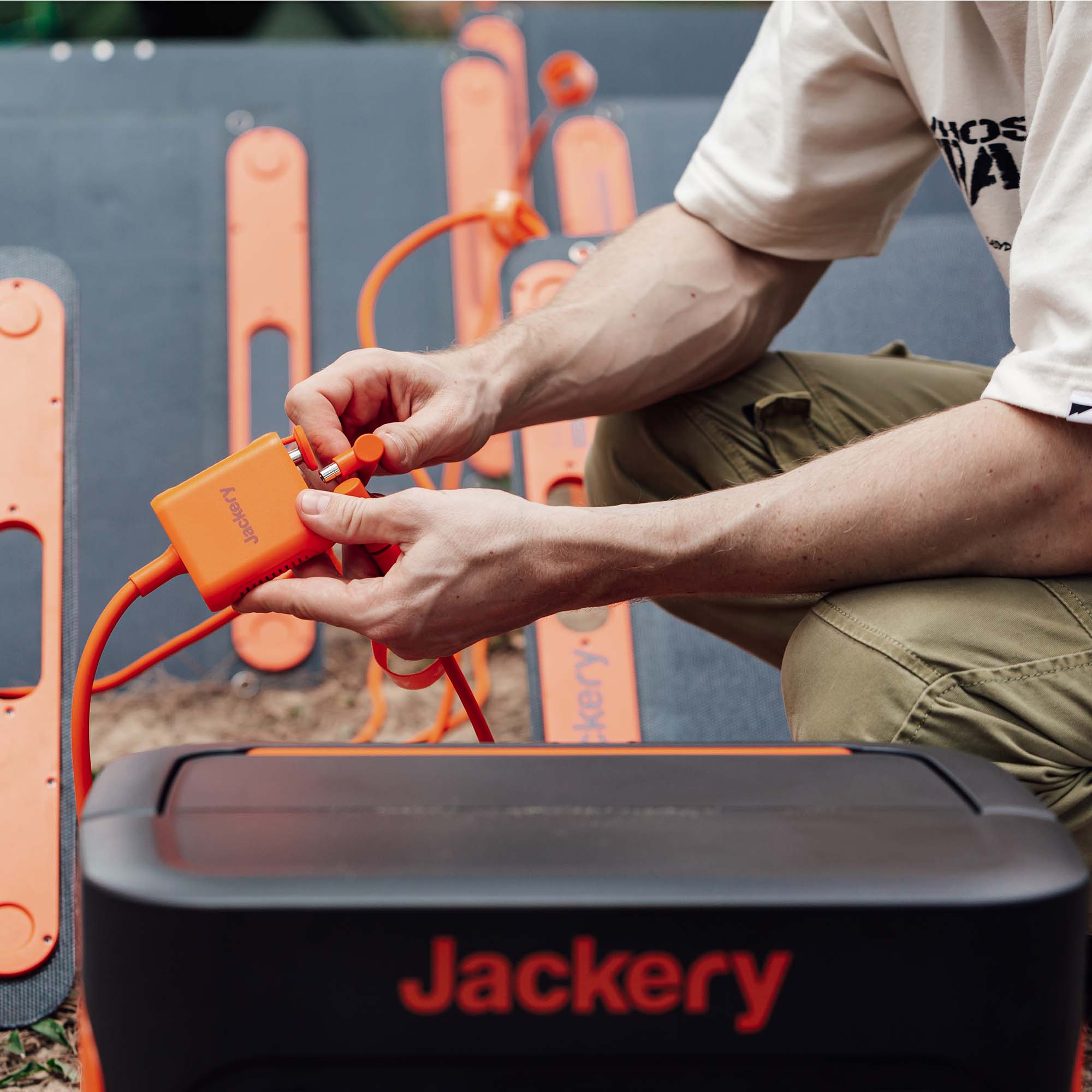 A user connects orange cables with a solar panel connector to a Jackery portable power station, surrounded by several orange Jackery solar panels. They wear olive green pants and a light-colored shirt.