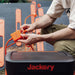 A user connects orange cables with a solar panel connector to a Jackery portable power station, surrounded by several orange Jackery solar panels. They wear olive green pants and a light-colored shirt.