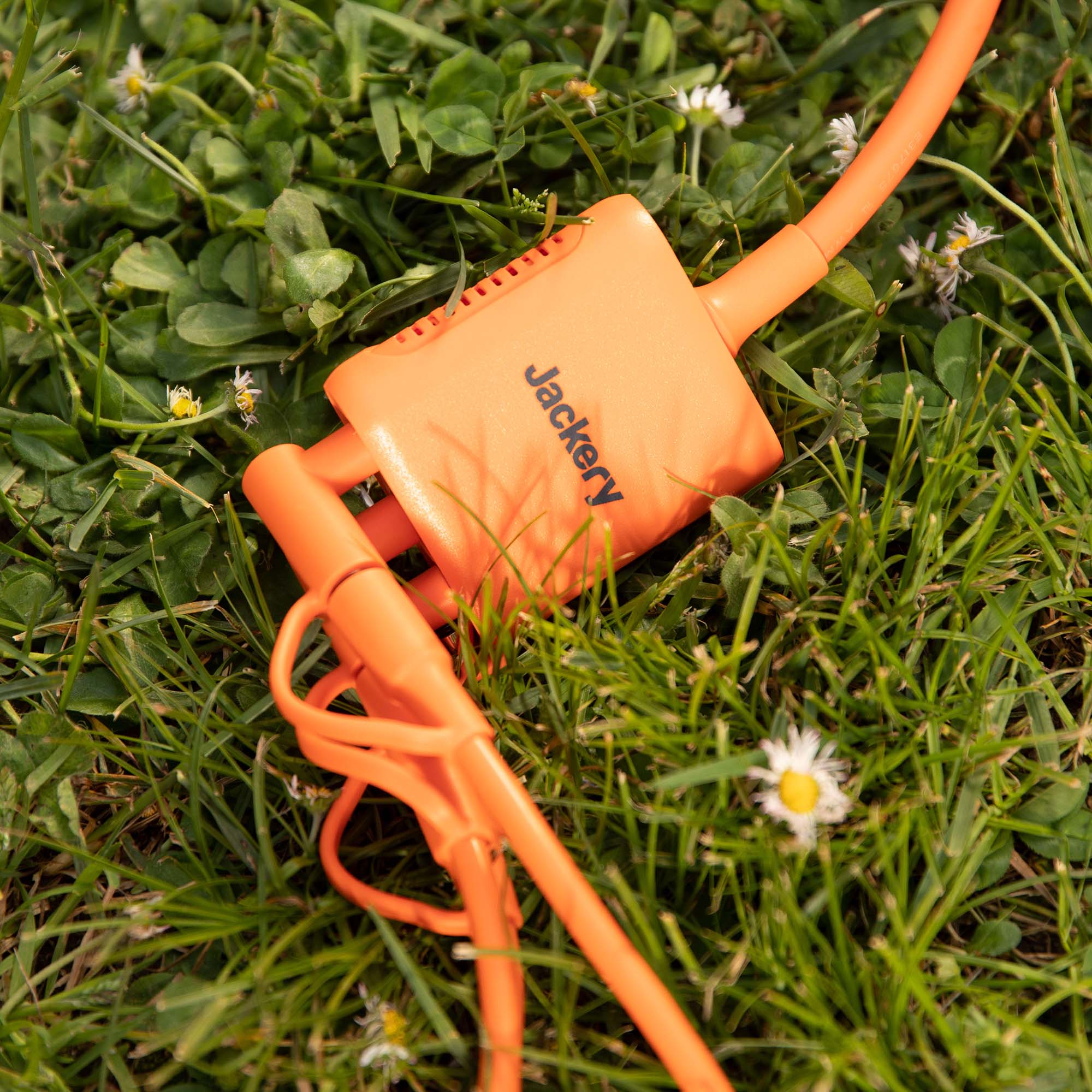 A Jackery-branded orange power cable and solar panel connector are placed on green grass dotted with small white flowers.