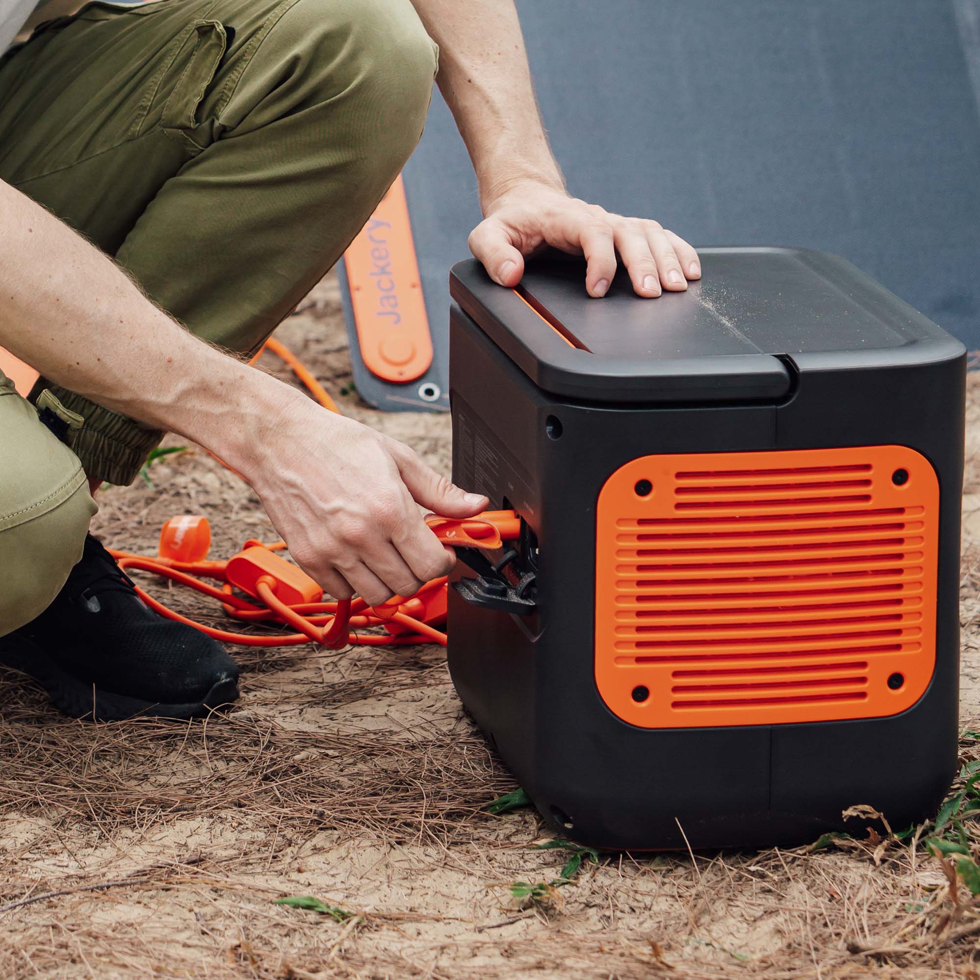 A person in green pants and black shoes connects a solar panel cable to a Jackery black and orange portable power station outdoors on dry grass.