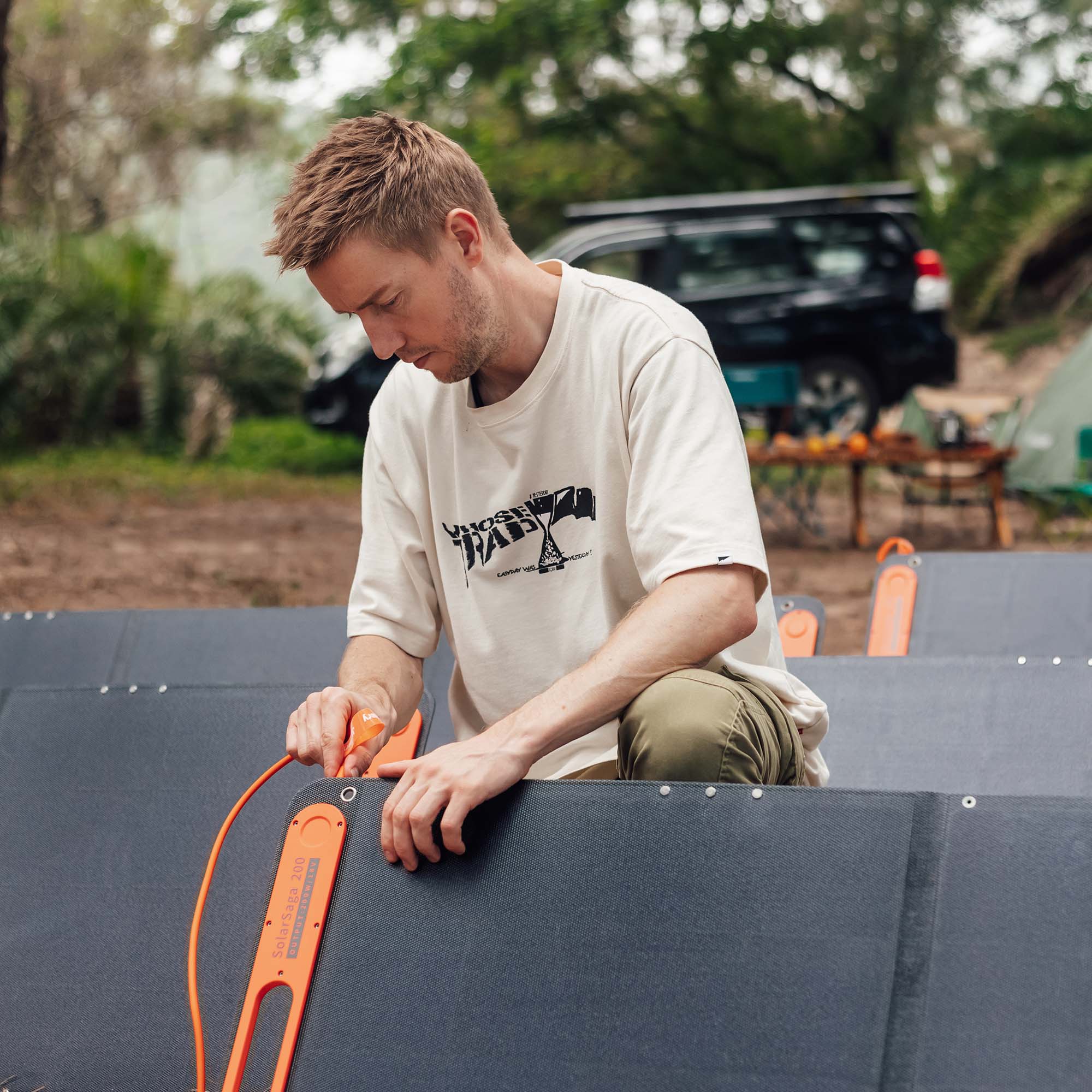 Outdoors, a person kneels and connects an orange Jackery solar extension cable to large black solar panels, with a parked SUV and campsite gear visible in the blurred background among trees.