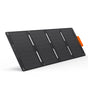A rectangular black Jackery portable solar charger with multiple small grid-like cells, four corner mounting holes, and an orange tab on one edge is shown standing at an angle against a plain white background.