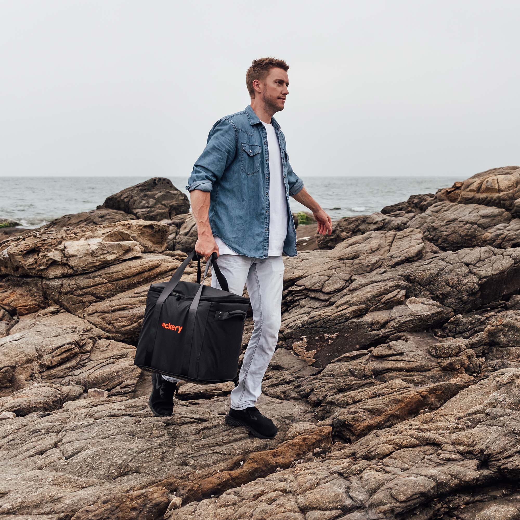 A man walks across rocky terrain near the ocean, wearing a light denim shirt and white pants. He carries a Jackery outdoor power station bag with "Catering" printed in red on the side.