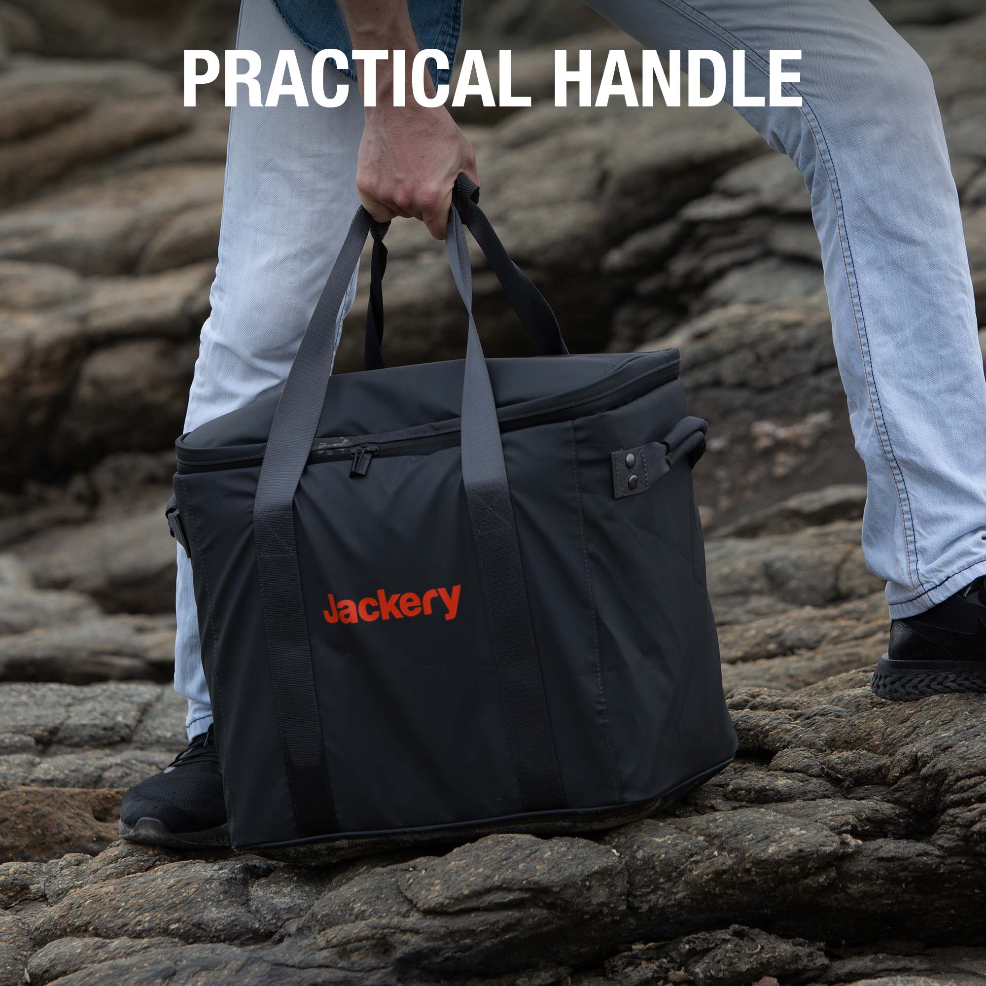 A person in jeans and black shoes carries a Jackery outdoor power station bag with two handles over rocky terrain. The text “PRACTICAL HANDLE” appears above, showing only the lower half of the person.