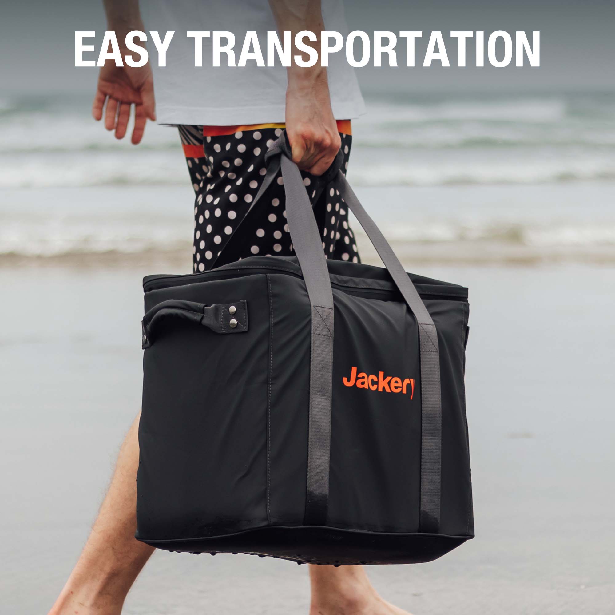 A person in patterned shorts carries a large black Jackery portable power station carrying case with gray straps on a beach. The text "EASY TRANSPORTATION" appears at the top.