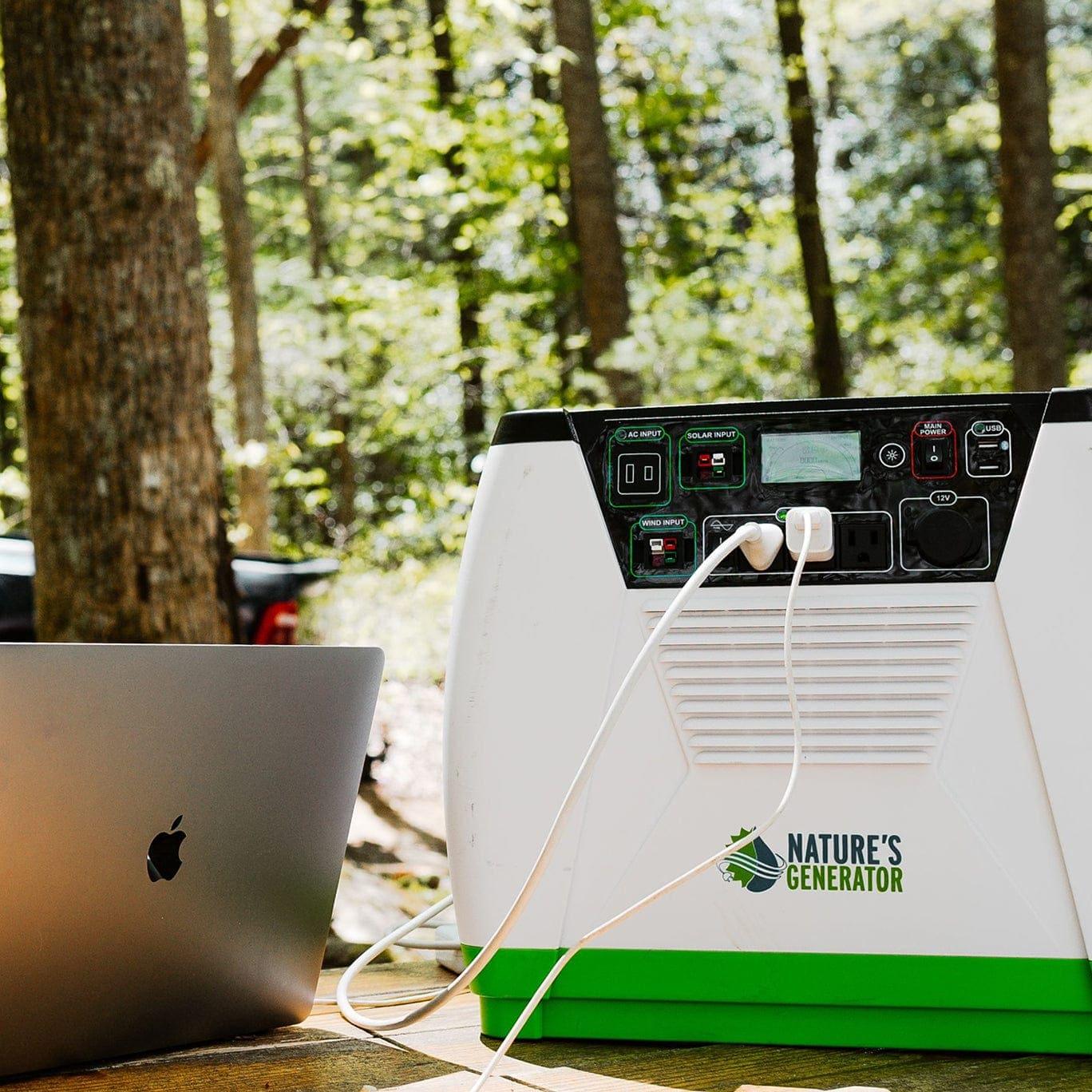 A Nature's Generator portable solar generator rests on a wooden surface outdoors, charging a laptop via two cables amid trees and sunlight in an off-grid forest setting.
