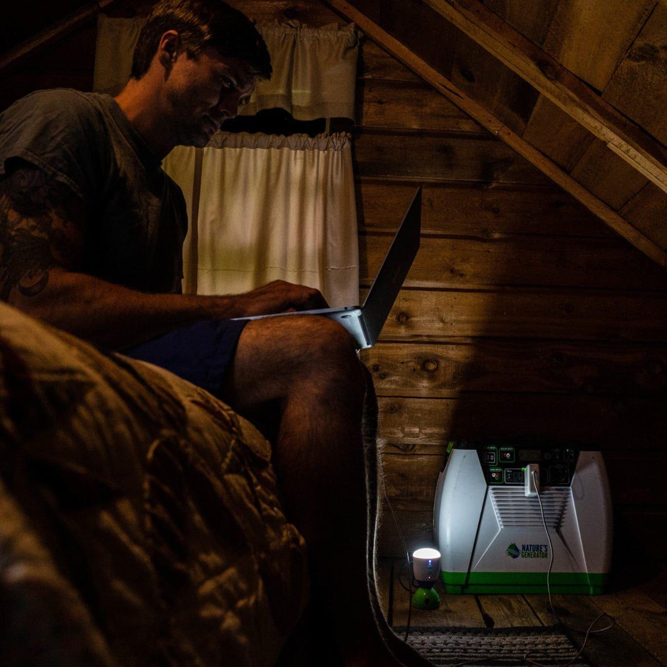 A person sits on a bed in a wooden cabin, using a laptop. Nearby on the floor, a Nature's Generator solar generator and small lantern supply renewable energy and light to the dimly lit room.