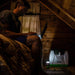 A person sits on a bed in a wooden cabin, using a laptop. Nearby on the floor, a Nature's Generator solar generator and small lantern supply renewable energy and light to the dimly lit room.