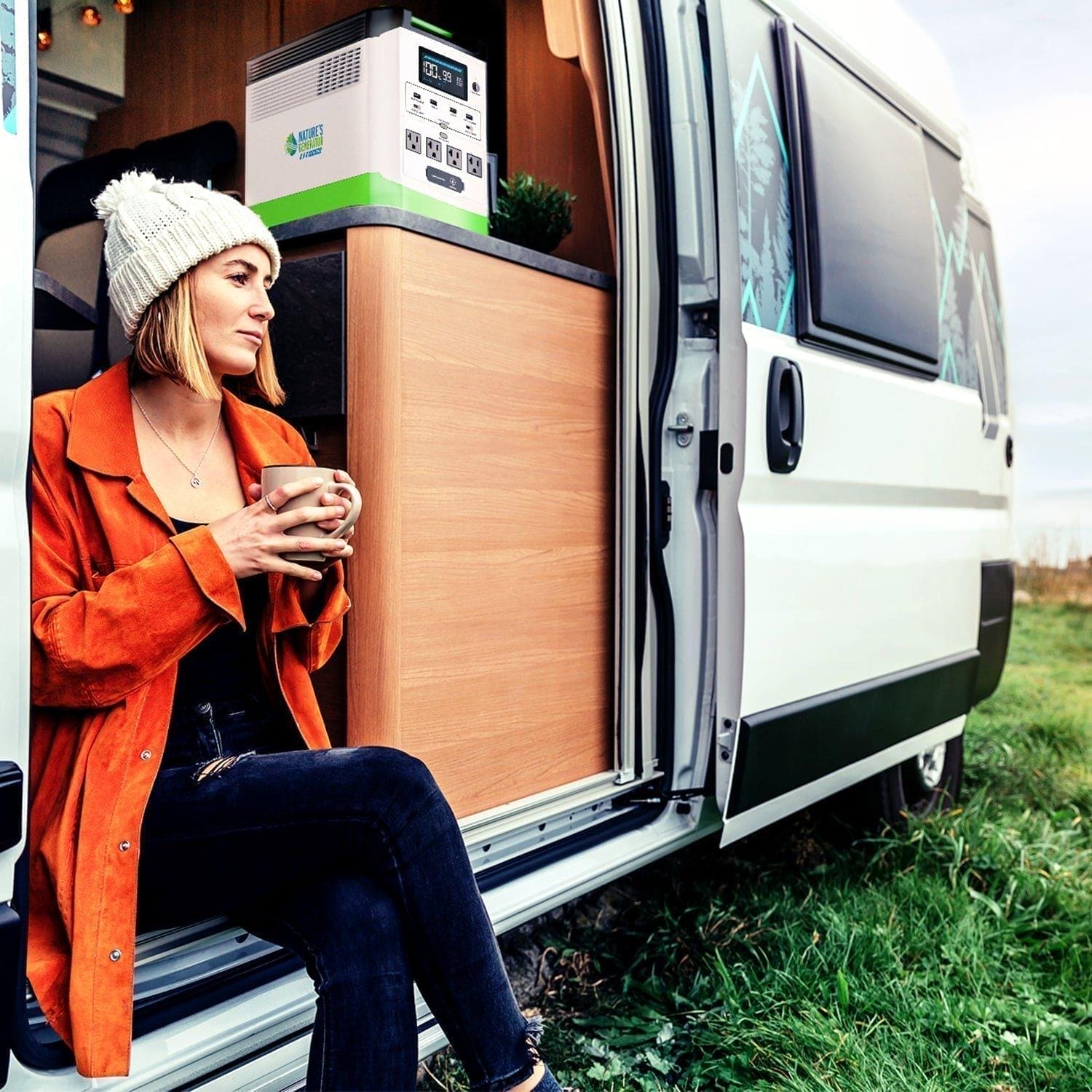 A woman in an orange jacket and white knit hat sits on the camper van’s step by grassy ground, holding a mug. Inside the open door is a Nature's Generator portable power station with LiFePO4 battery. She appears relaxed.