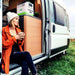 A woman in an orange jacket and white knit hat sits on the camper van’s step by grassy ground, holding a mug. Inside the open door is a Nature's Generator portable power station with LiFePO4 battery. She appears relaxed.