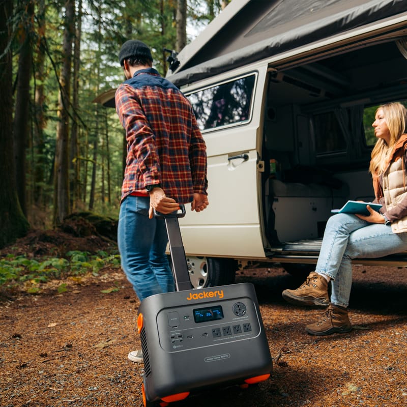 A person drags a high-capacity Jackery power station using a handle close to a white camper van surrounded by trees, as another individual sits at the van's doorway with a notebook in hand. Both are dressed casually for outdoor activities, preparing to enjoy the Jackery's convenient portability that complements their adventurous lifestyle.