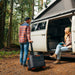 A man and a woman are next to a camper van in a forest setting. The man is holding a high-capacity portable power station on wheels by Jackery. The woman is seated at the entrance of the van, holding a notebook. The open pop-up roof of the van merges effortlessly with the trees around it.