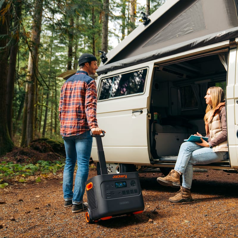 A man and a woman are next to a camper van in a forest setting. The man is holding a high-capacity portable power station on wheels by Jackery. The woman is seated at the entrance of the van, holding a notebook. The open pop-up roof of the van merges effortlessly with the trees around it.