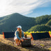 Dressed in outdoor gear, a person kneels beside the Jackery portable solar power generator, renowned for its large capacity, in a picturesque mountain setting. Two solar panels lie on the ground nearby. The sky is mostly clear with some clouds, and lush pine forests can be seen in the distance.