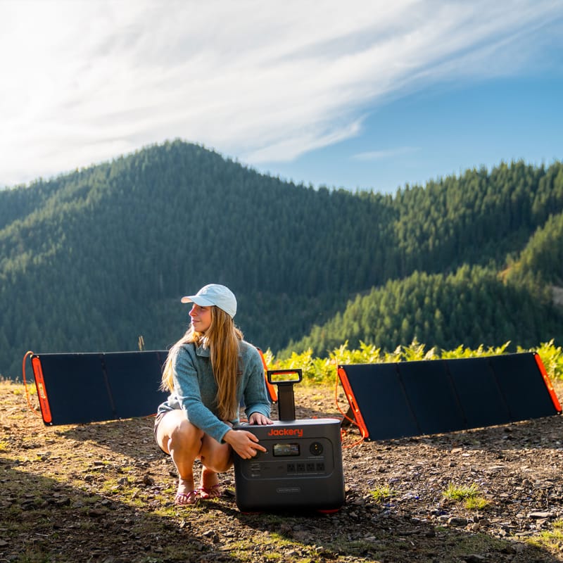 Dressed in outdoor gear, a person kneels beside the Jackery portable solar power generator, renowned for its large capacity, in a picturesque mountain setting. Two solar panels lie on the ground nearby. The sky is mostly clear with some clouds, and lush pine forests can be seen in the distance.