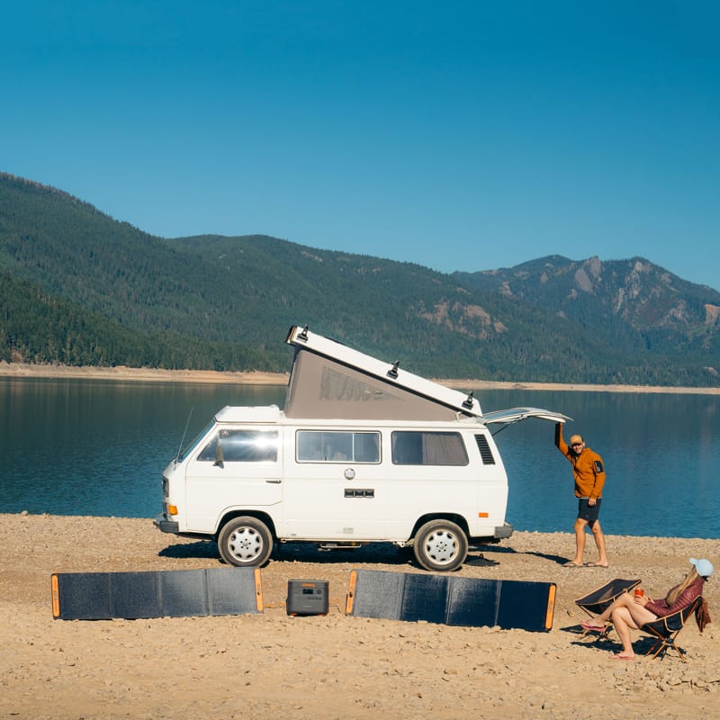 A Jackery camper van with an open roof and spacious interior is parked by a lake, with majestic mountains in the background. One person stands near the van while another lounges on a camping chair. Jackery solar panels are positioned on the ground, providing ultra-fast charging amid nature's splendor.