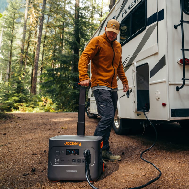 A person wearing a brown jacket and cap plugs the high-capacity Jackery Portable Power Station into a motorhome surrounded by trees and lush greenery, creating a serene outdoor camping scene.