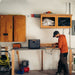 A person in an orange hoodie works on an electronic device at a wooden workbench in a garage. A Jackery portable power station with ultra-fast charging capabilities sits on the bench, surrounded by various tools and equipment.