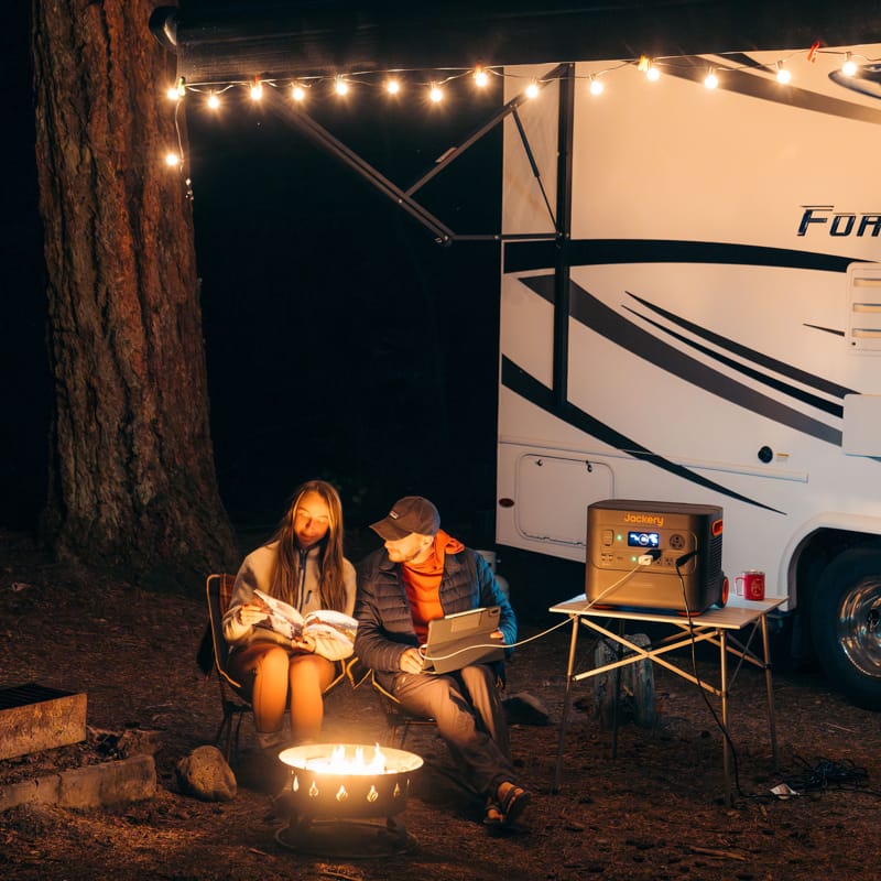 Two people relax by a campfire under the night sky; one engrossed in a book while the other uses a ultra-fast charging Jackery laptop. They are beside a white camper adorned with string lights, accompanied by a small table holding a portable Jackery refrigerator. Tall trees encircle the serene campsite.